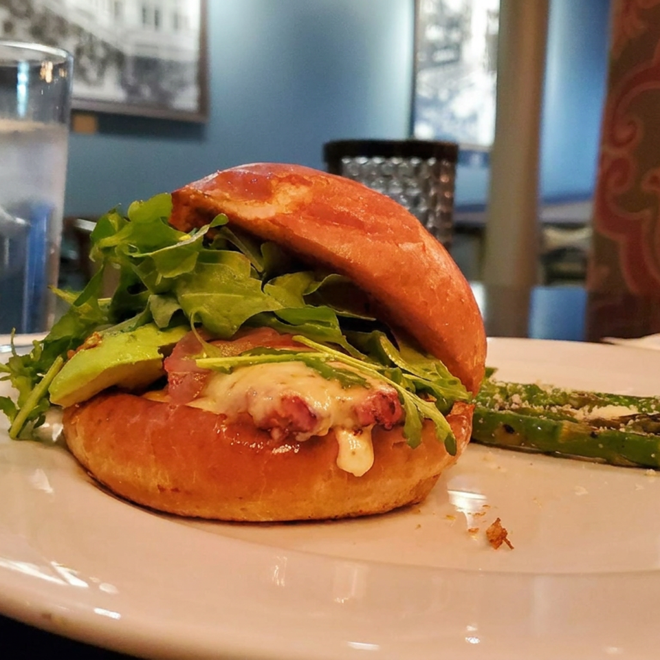 Gourmet burger with arugula, avocado, melted cheese, and sauce served alongside grilled asparagus on a white plate.