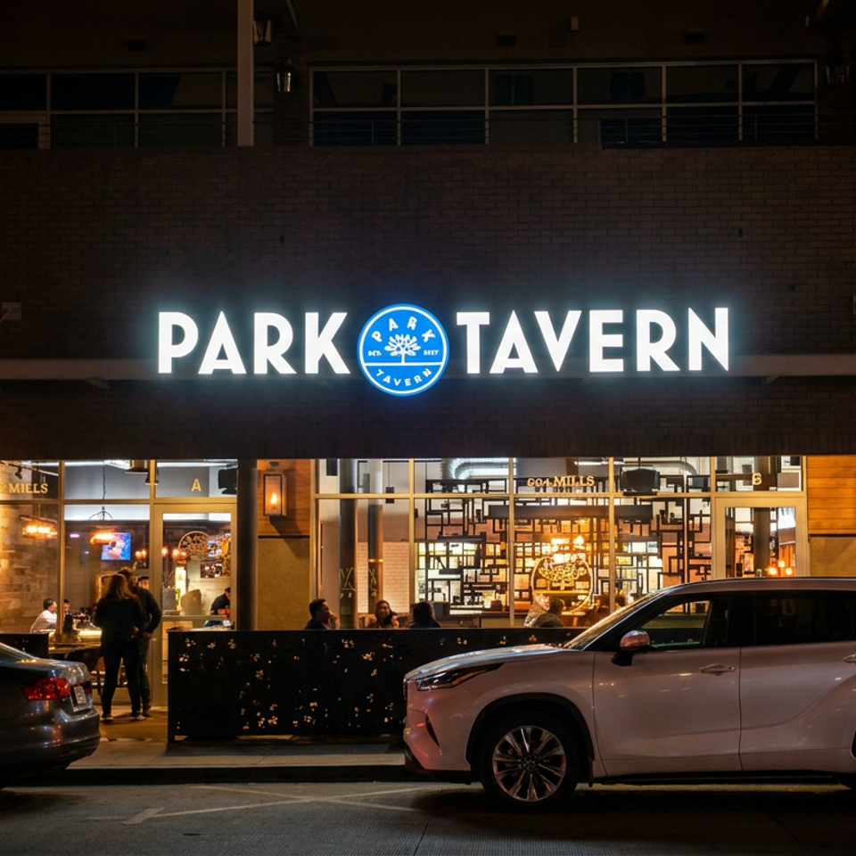 Park Tavern restaurant entrance glowing at night, with illuminated signage, outdoor seating, diners, and parked cars.