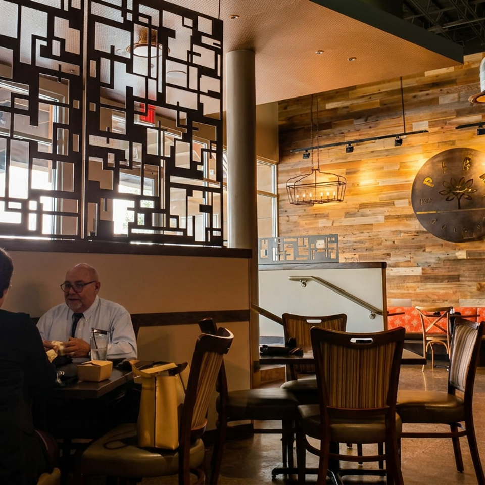 Warmly lit restaurant dining room with three people seated at a table, geometric partition, wood-paneled walls, and pendant lights.