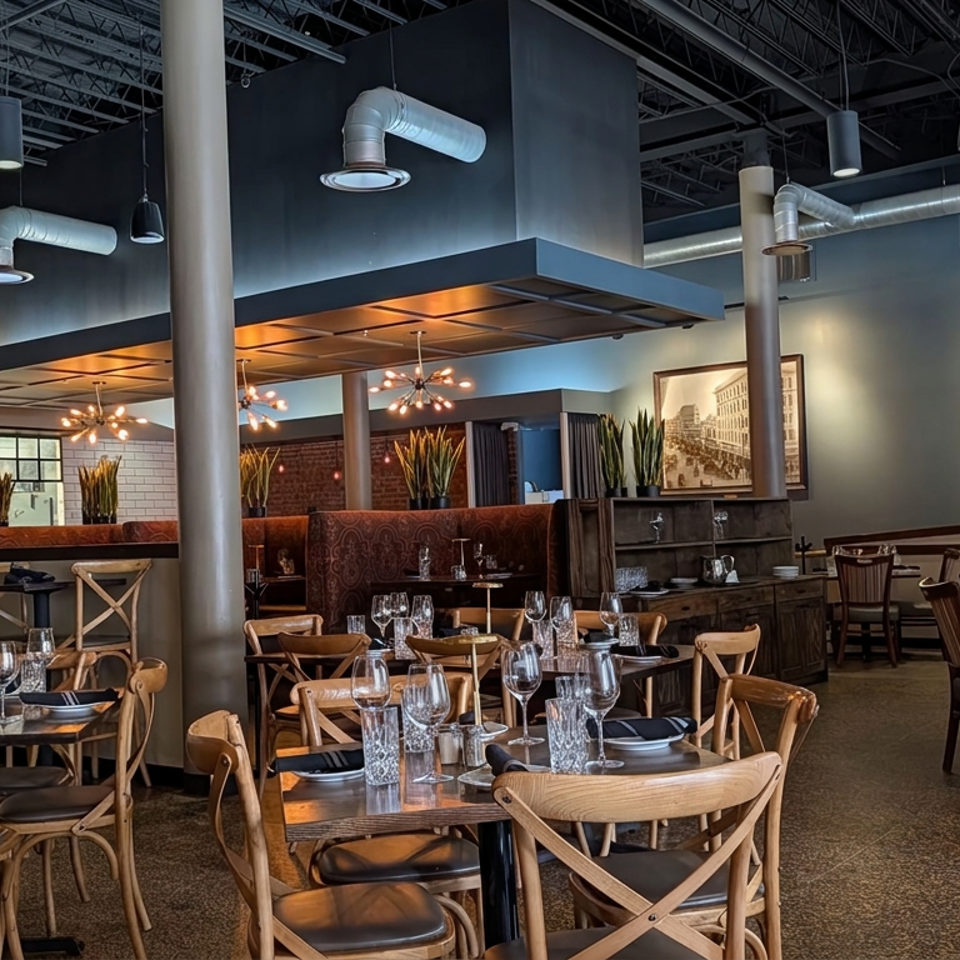 Modern industrial restaurant dining room with set tables, wooden chairs, exposed ductwork, booth seating, and framed artwork.